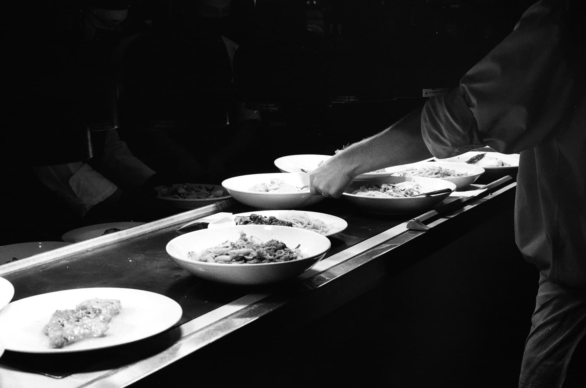 Plates of food lined up on a dark kitchen pass as a chef reaches across