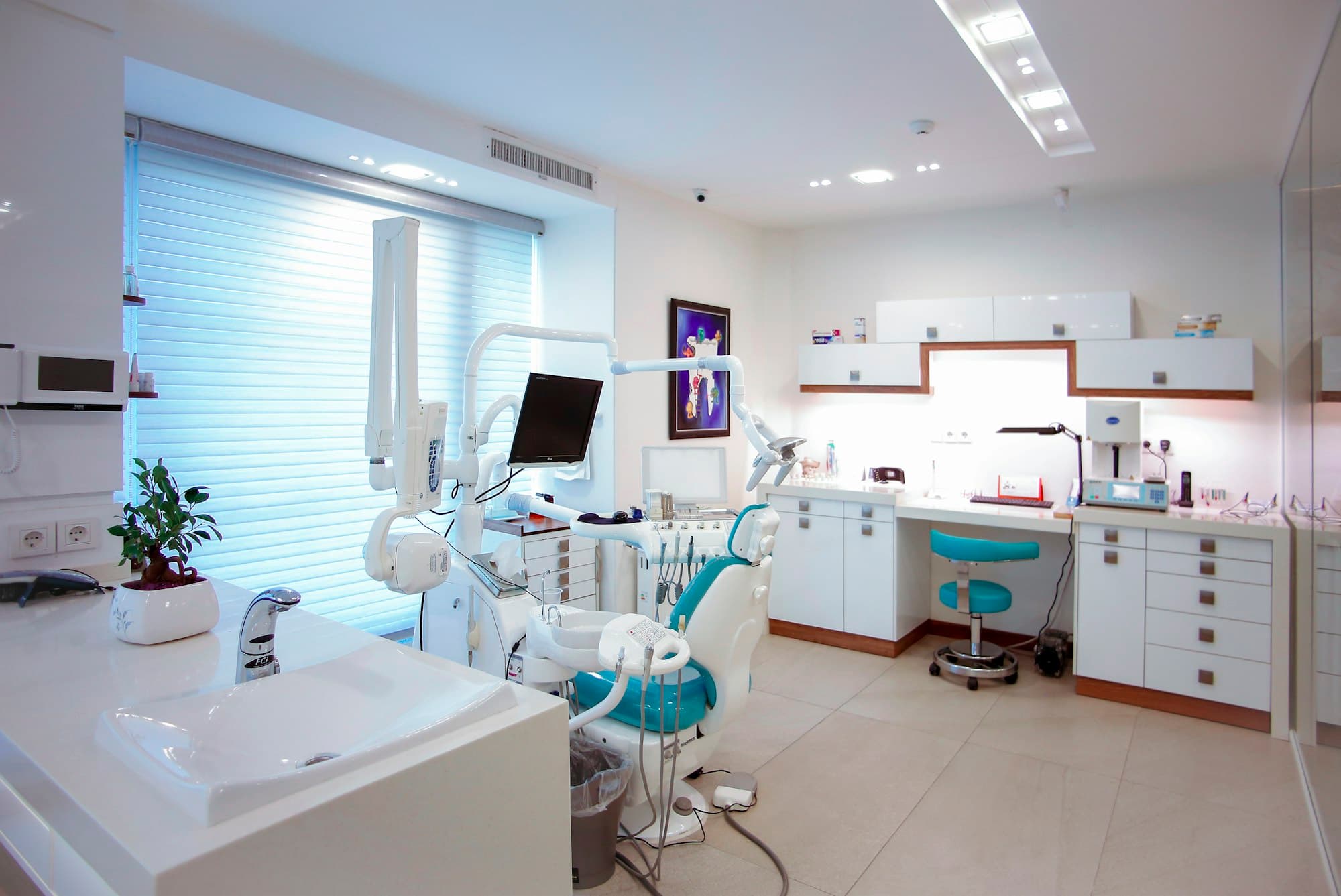 A modern dental treatment room with a teal chair, wall-mounted screen and daylight through the blinds